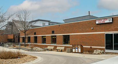 An exterior photo of the Student Union Building at Fanshawe College. A sign states, Student Union Building. The Out Back Shack logo is displayed.