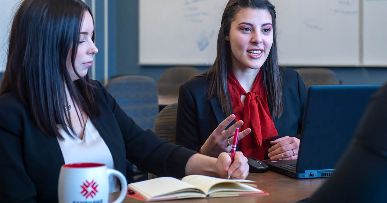 Two young women engaged in a discussion during a meeting, one taking notes in a notebook while the other speaks, with a laptop open on the table.