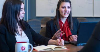 Two young women engaged in a discussion during a meeting, one taking notes in a notebook while the other speaks, with a laptop open on the table.
