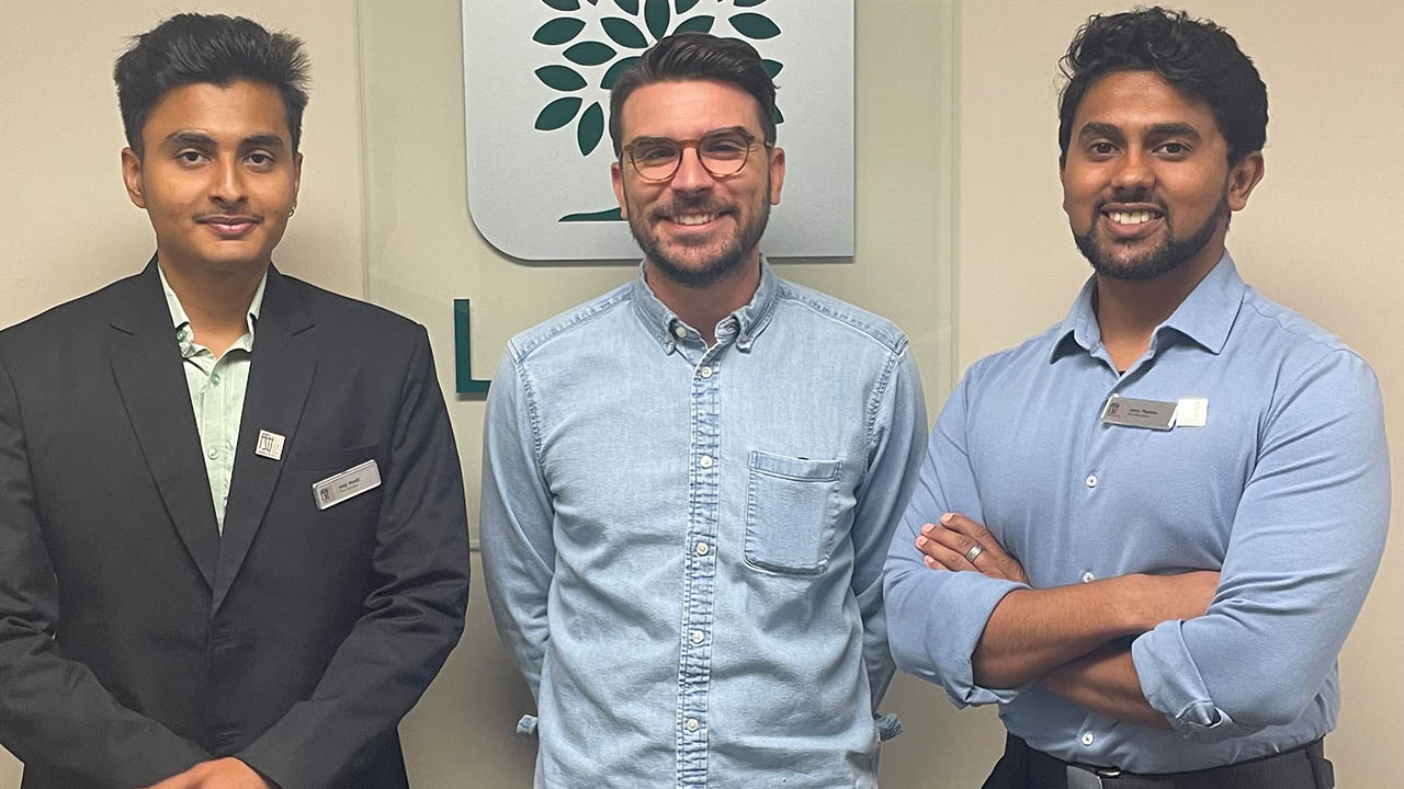 Three men standing indoors in front of a wall with a green tree logo, dressed in business casual and formal attire.