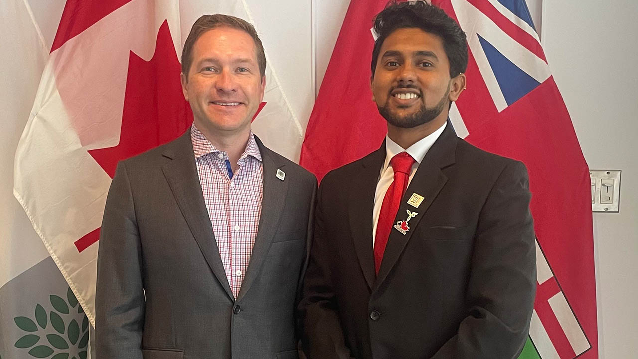 Two men in formal suits stand side by side in front of Canadian and Ontario flags indoors.