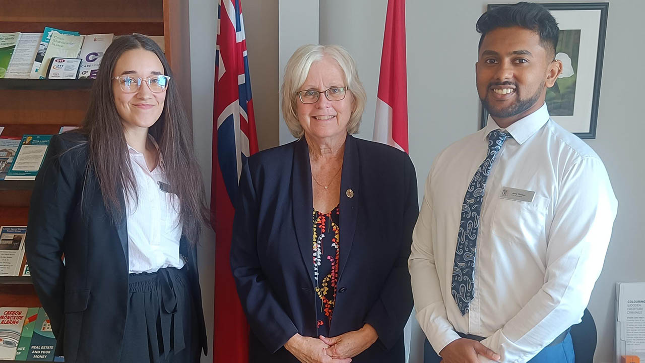 Three professionals stand indoors with two flags behind them, dressed in formal business attire including blazers and a tie.