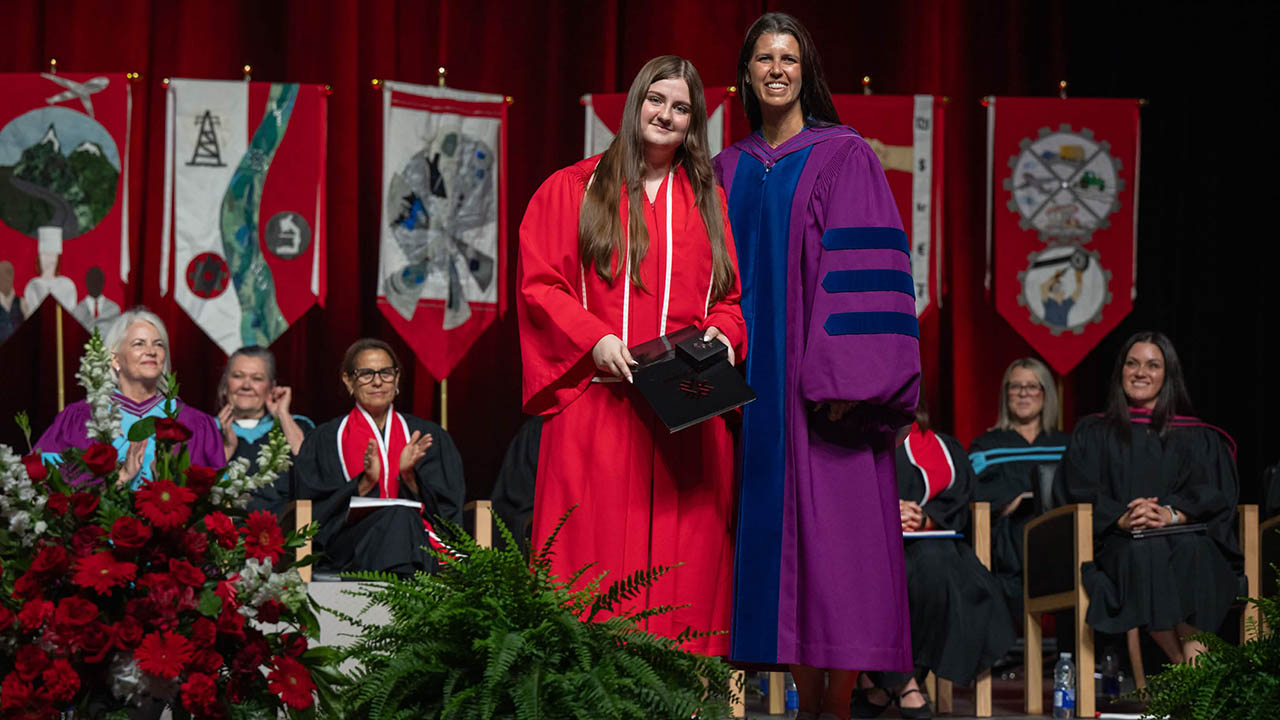 Two women smiling on stage during a graduation ceremony, one in a red cap and gown holding a diploma cover, the other in a purple academic robe.