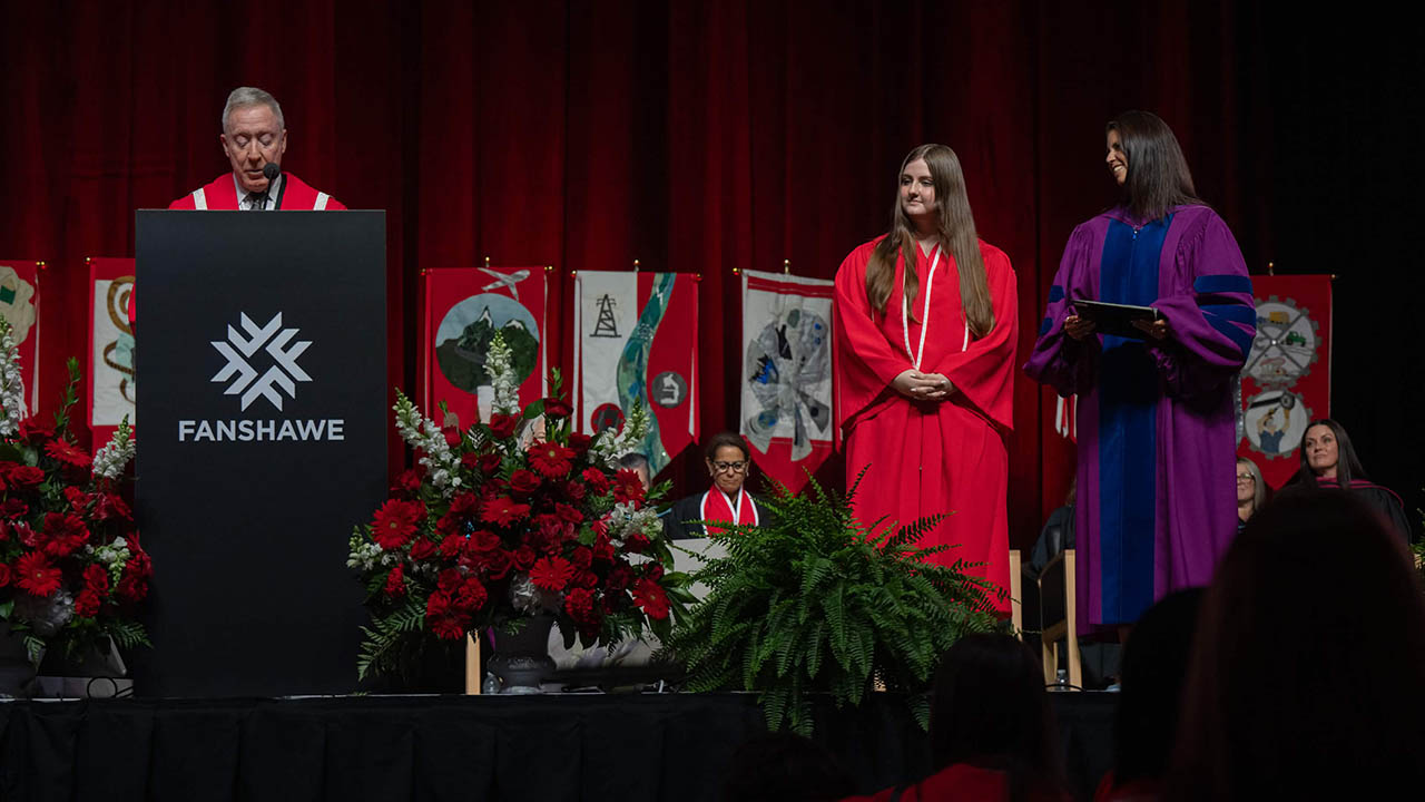 A graduate in a red gown stands on stage smiling beside a faculty member in purple robes during a Fanshawe College ceremony.