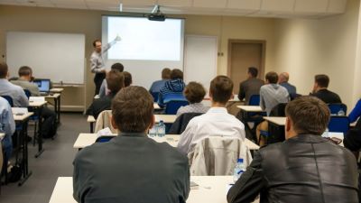 Students attentively listen to a lecturer presenting information on a projector screen in a classroom setting.