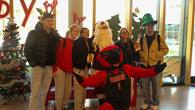 Group of people posing indoors with a person dressed as Santa and a red mascot in a basketball jersey, festive decorations in the background.