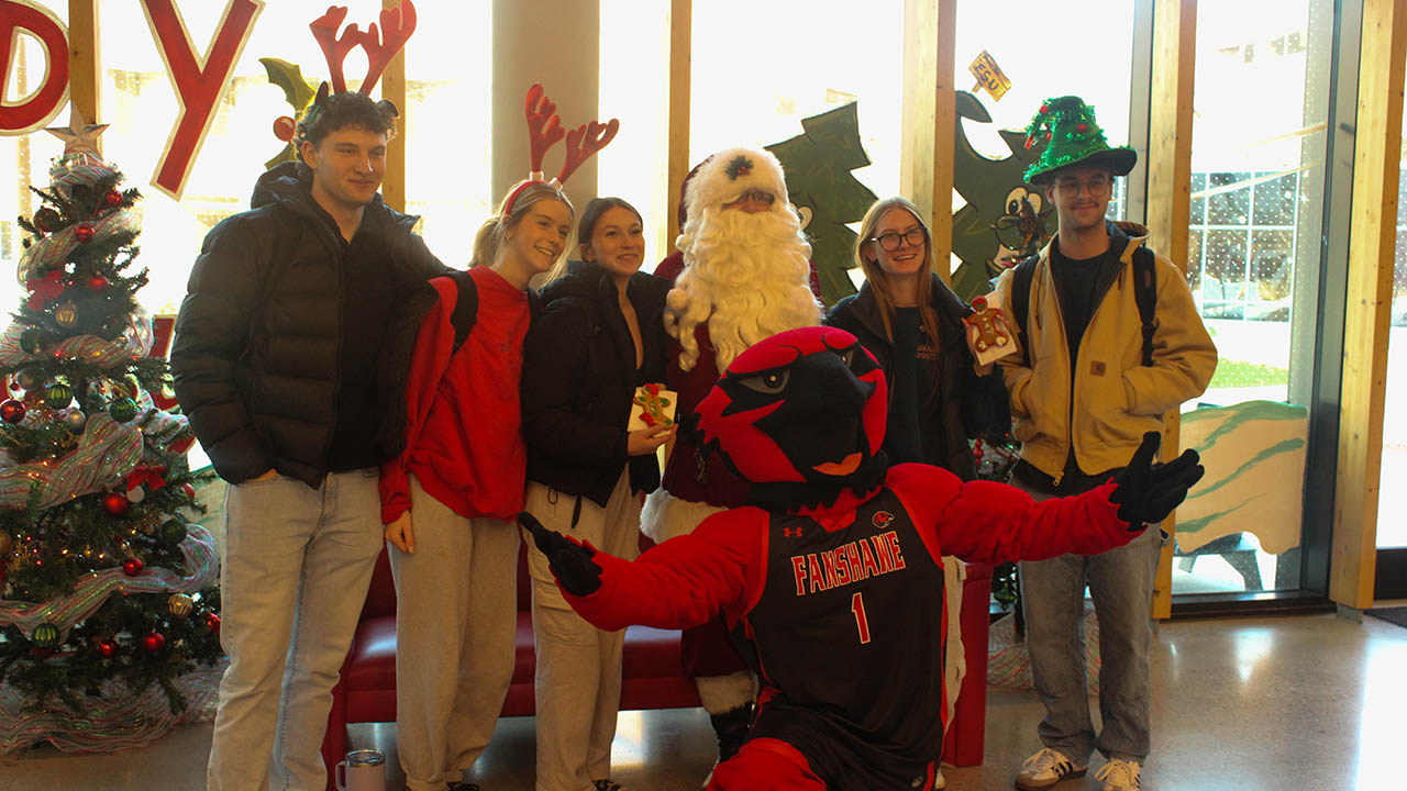 Group of people posing indoors with a person dressed as Santa and a red mascot in a basketball jersey, festive decorations in the background.
